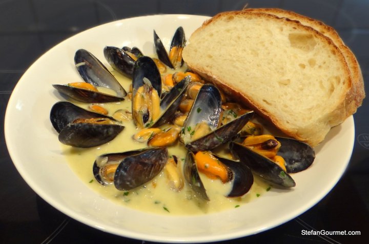 A bowl of mussels in a creamy sauce, garnished with herbs, accompanied by a slice of bread.