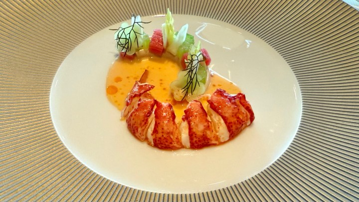 A beautifully plated dish featuring lobster tail with a sauce and a side of colorful garnishes, including green vegetables and small cubes of red fruit.