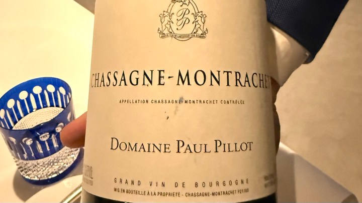 A close-up of a wine bottle labeled 'Chassagne-Montrachet' from Domaine Paul Pillot, accompanied by a glass of water in a blue decorative glass.