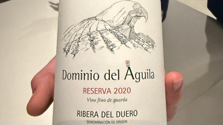 A person holding a bottle of Domínio del Águila Reserva 2020 wine from Ribera del Duero, featuring an illustrated label with a vineyard design.