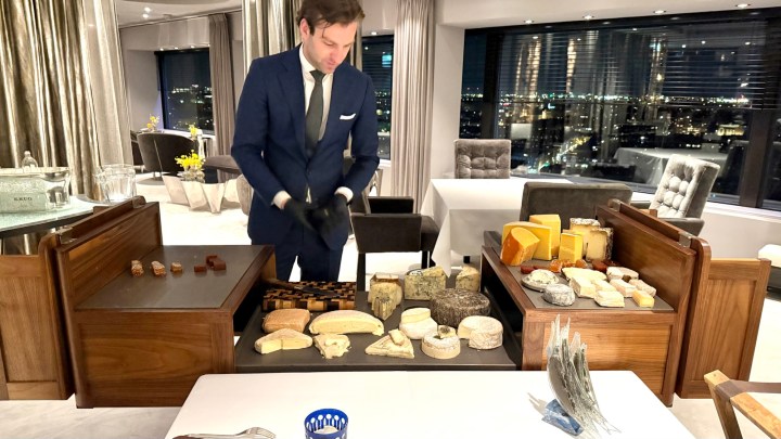 A waiter in a formal outfit is presenting a selection of various cheeses on a wooden cart, with a city view visible through large windows in the background.