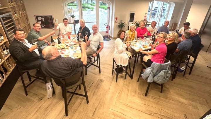A group of people gathered around a dining table, enjoying a meal and raising their glasses in a toast, with a modern vineyard lounge in the background.