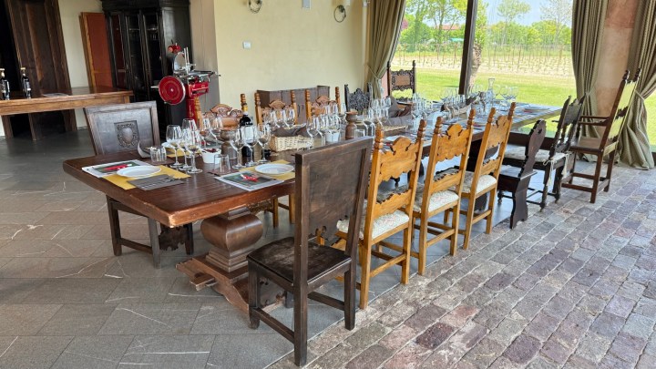 A spacious dining area with a long wooden table set for a wine tasting, featuring glasses and various tasting notes, overlooking a vineyard through large windows.