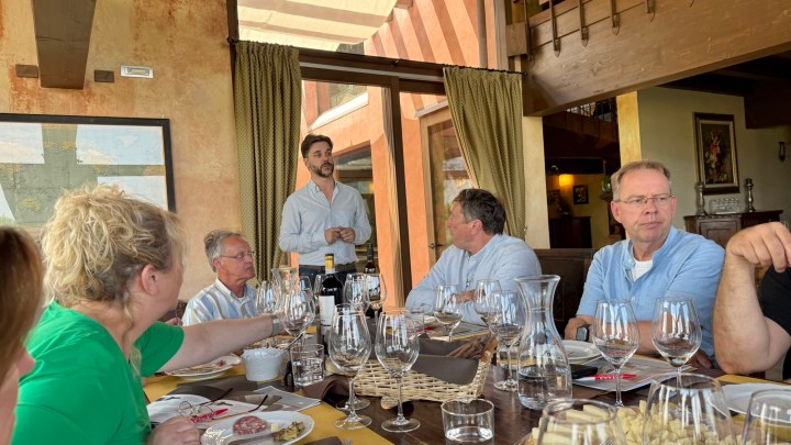 A group of people sitting around a dining table with wine glasses, engaged in conversation while a man stands and speaks in front of a large window.