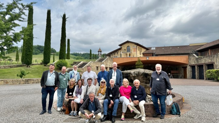 A group of wine enthusiasts gathered outside a winery in Italy, with a scenic backdrop of trees and a building. They are smiling and posing for the photo.