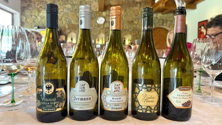Five wine bottles on a table with glasses and a clock in the background. The bottles include 'Vinnae Ribolla Gialla 2024', 'Jermann Pinot Grigio 2024', 'Ramata Pinot Grigio 2022', 'Vintage Tunina', and 'Pignolo 2018', showcasing a variety of wines.