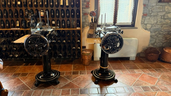Two antique meat slicing machines displayed in a wine cellar, with shelves full of wine bottles in the background.