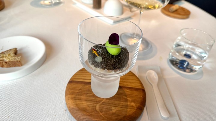 A glass dish containing caviar garnished with a green element and flower petals, placed on a wooden serving board, with a piece of bread and other dining items visible in the background.