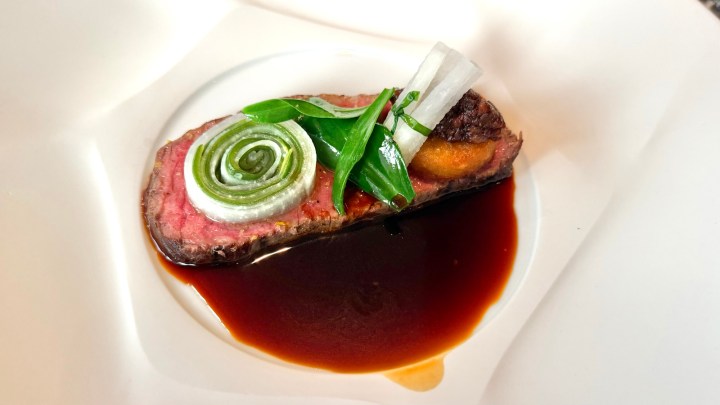 A beautifully plated dish featuring a slice of beef topped with green onions and a decorative swirl, served with a dark sauce.