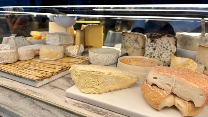 An assortment of cheeses displayed in a glass-fronted case, featuring various types including soft, hard, and blue cheeses.