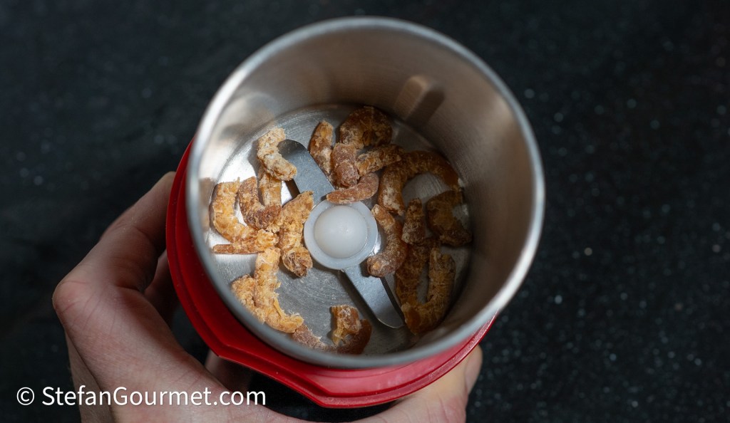 A person holding a spice grinder with dried shrimp pieces inside, ready to be ground into a powder.