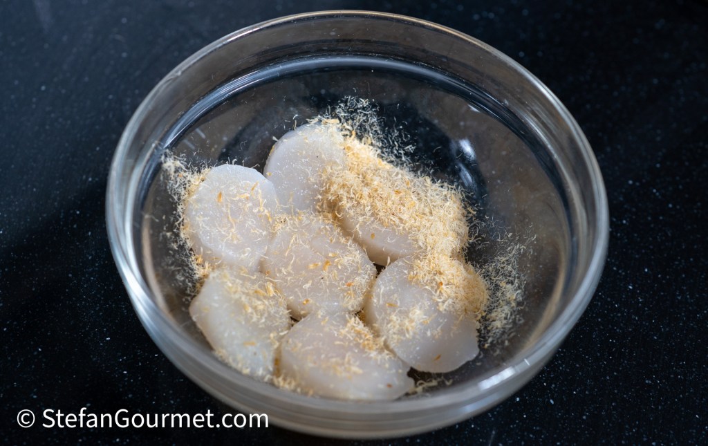 A bowl containing raw scallops topped with ground dried shrimp on a black surface.