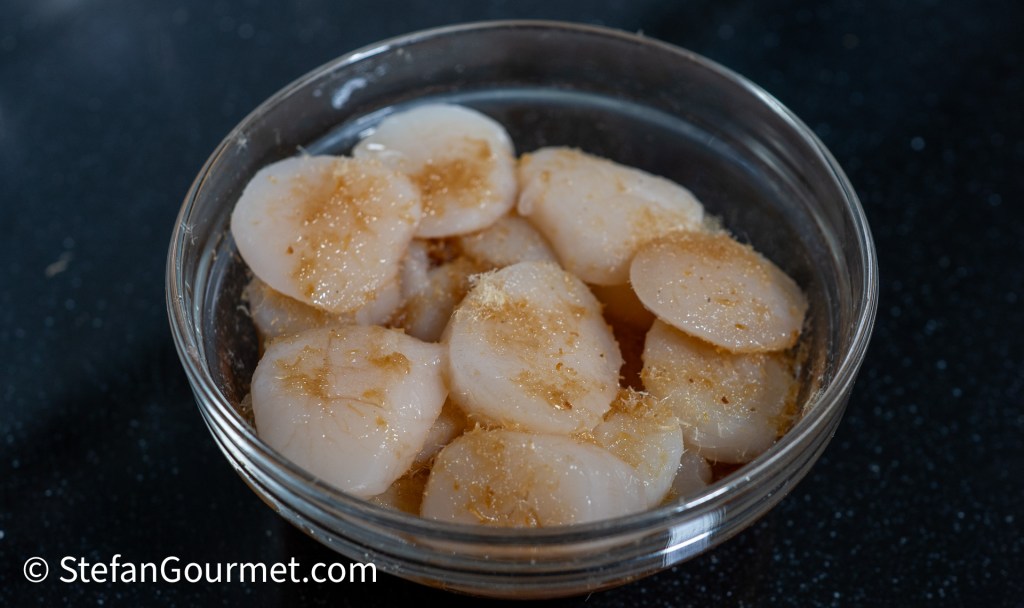 Bowl of marinated scallop slices topped with dried shrimp, ready for a seafood dish.