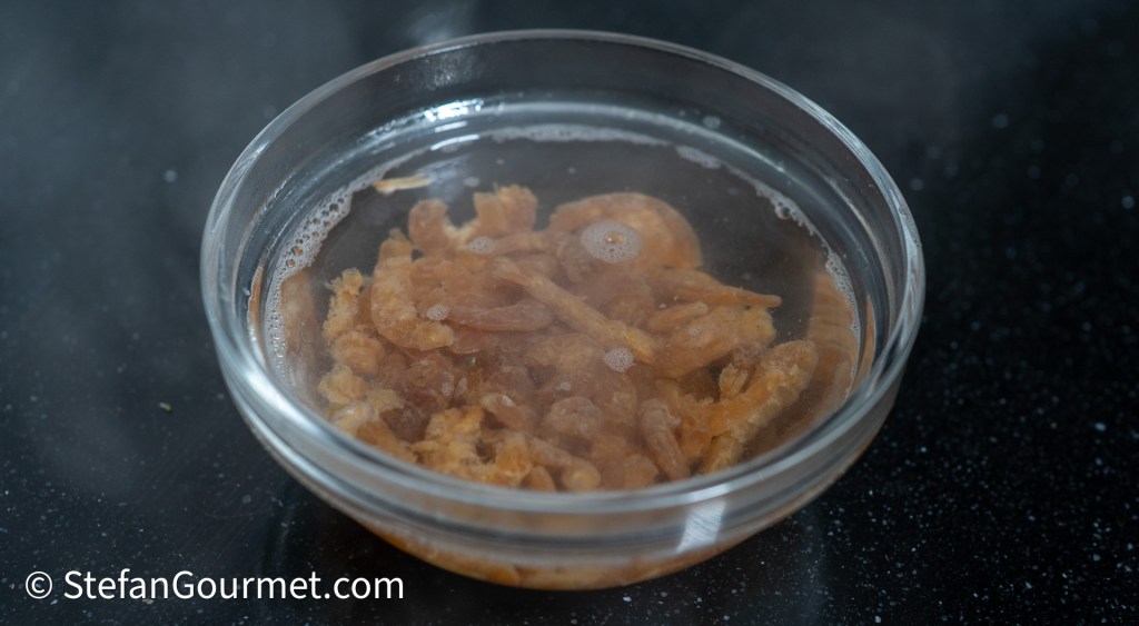 A bowl containing soaked dried shrimp in hot water, releasing steam, placed on a dark countertop.