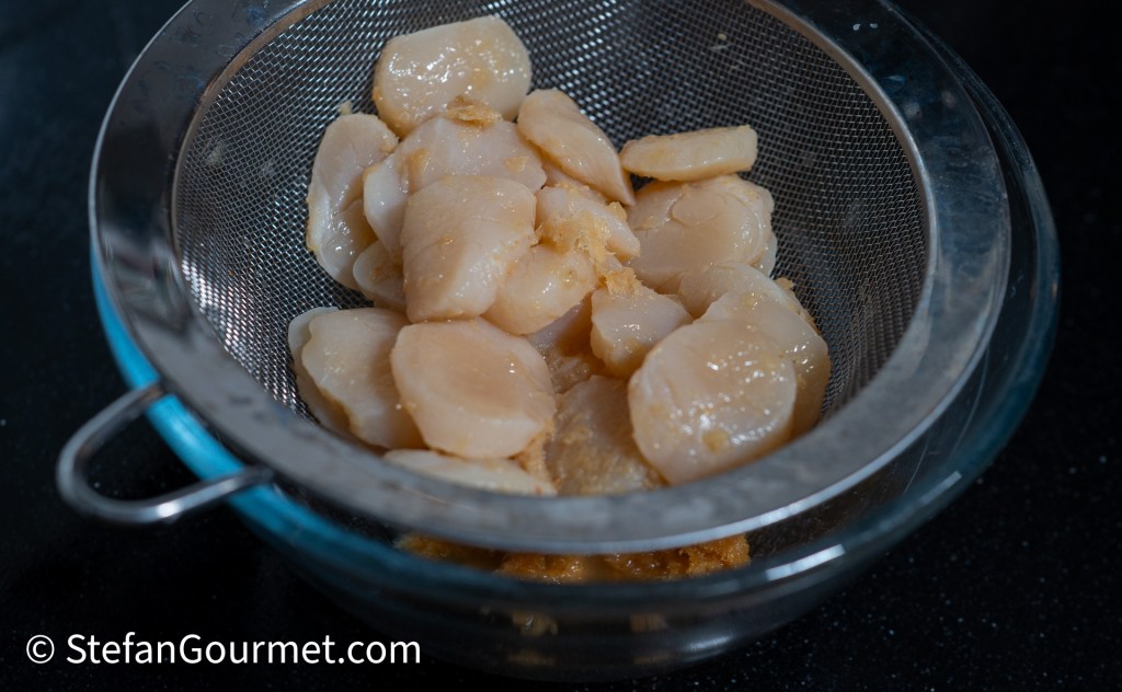Scallops draining in a fine mesh strainer over a glass bowl.