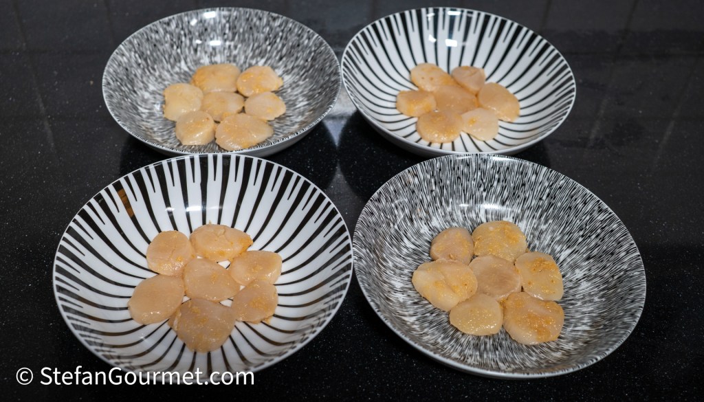 Four bowls with slices of raw scallops arranged in a flower pattern, resting on a black countertop.