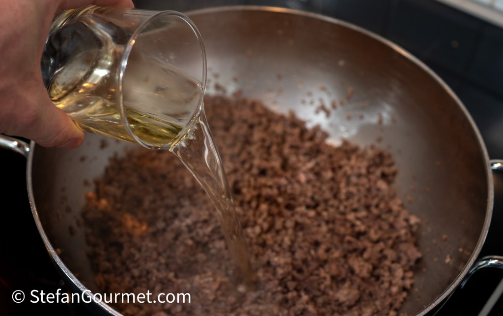 A close-up of a hand pouring dry white wine into a pan with browned ground beef, showing the meat mixture on the stove.