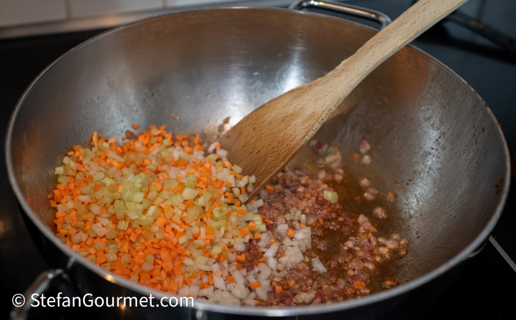 A sauté pan with finely chopped onions, carrots, and celery being stirred with a wooden spoon, with pieces of cooked pancetta at the bottom.