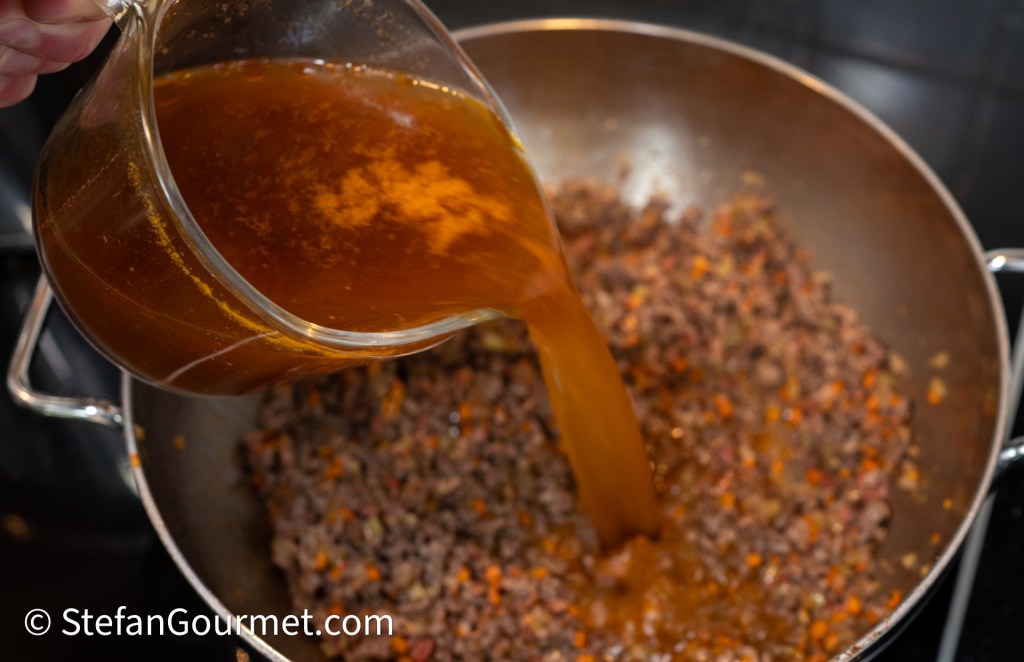A person pouring meat stock into a pan containing cooked ground meat and vegetables, showcasing the process of making a rich sauce.