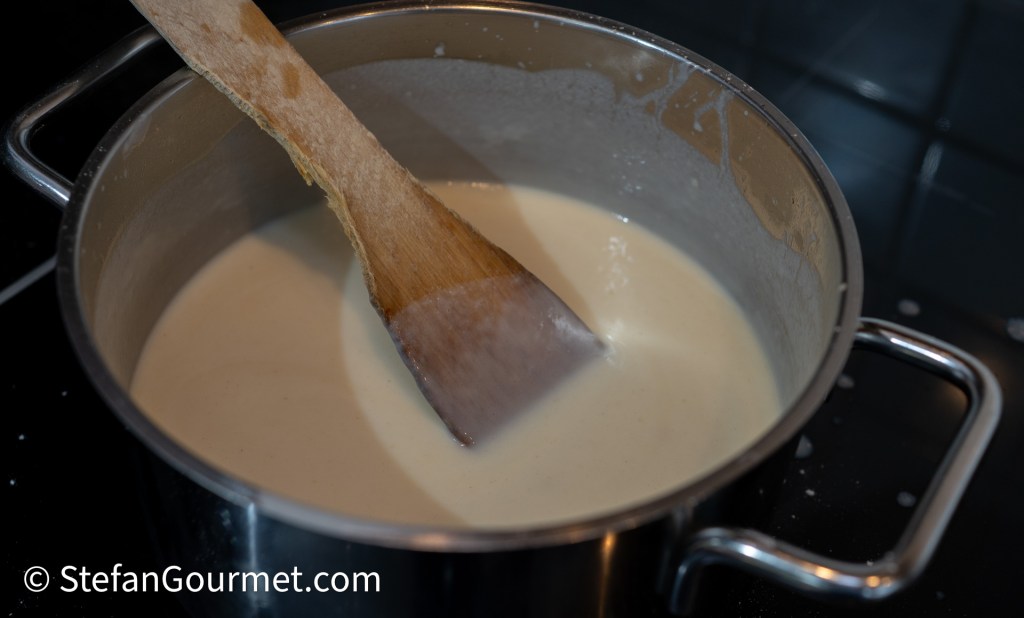 A saucepan containing a creamy white sauce, with a wooden spoon stirring it on a stovetop.