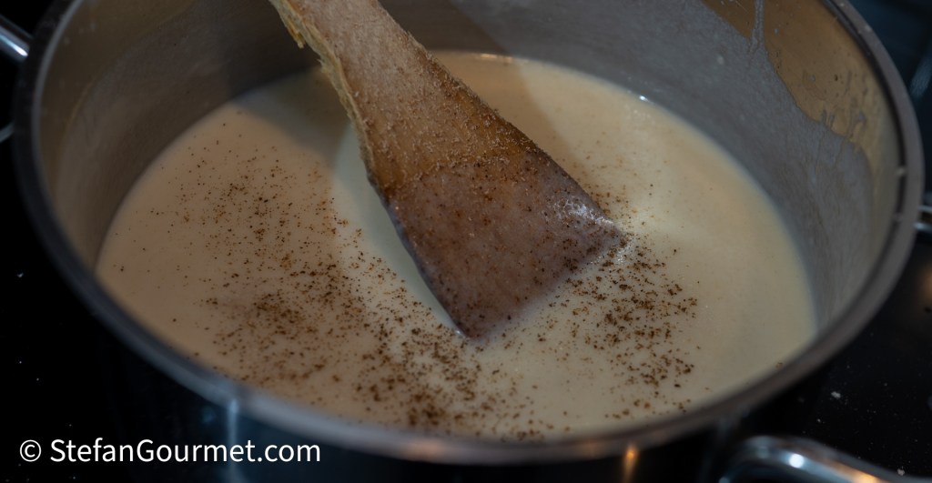 A pot of creamy white sauce with a wooden spoon stirring, topped with a dash of grated nutmeg.