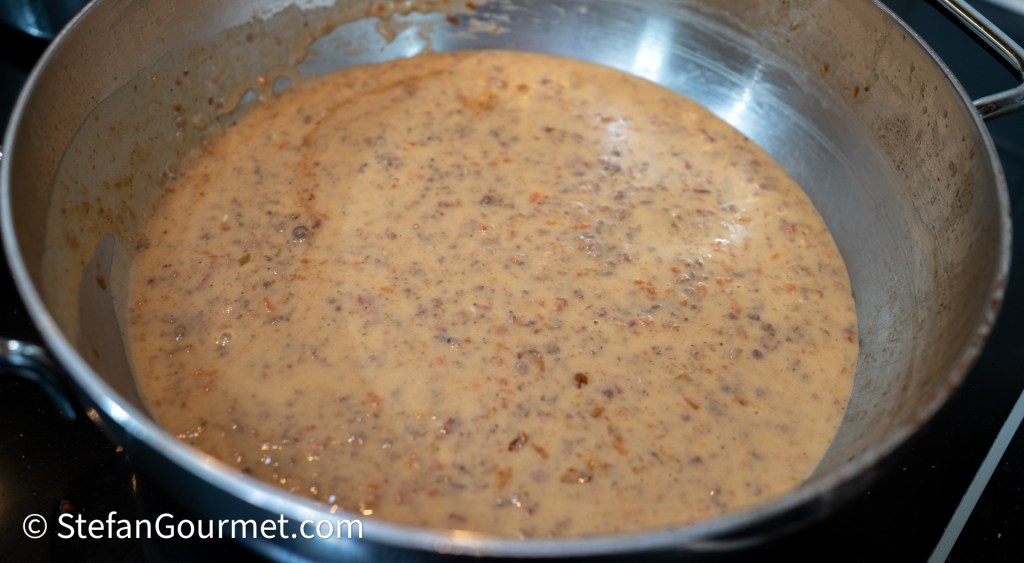A stainless steel pan filled with creamy meat ragù, showcasing a thick, rich sauce with ground meats and vegetables, cooking on a stovetop.
