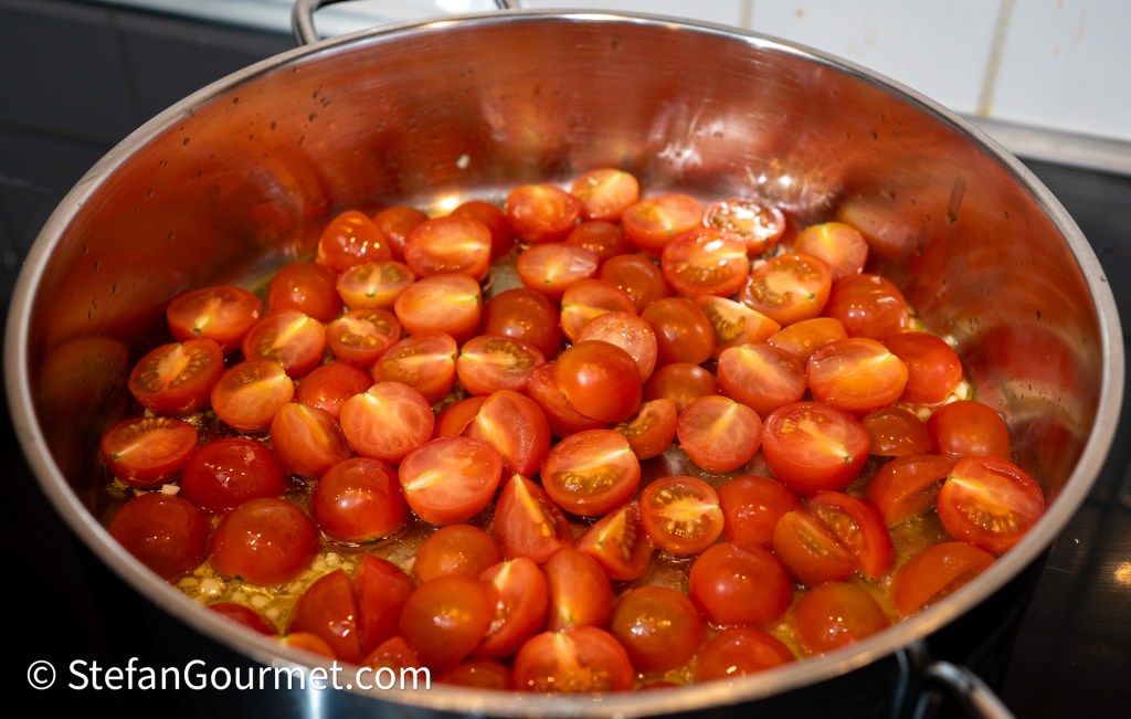 A stainless steel pan filled with halved cherry tomatoes cooking in oil and garlic, ready to be incorporated into a dish.