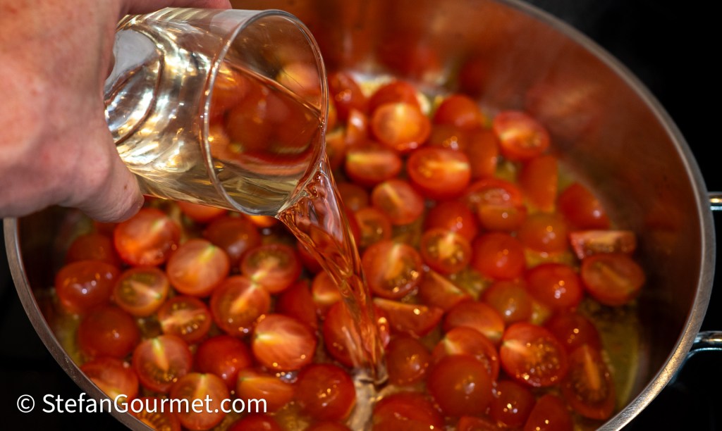 A hand pouring white wine into a pot filled with halved cherry tomatoes, cooking on the stove.