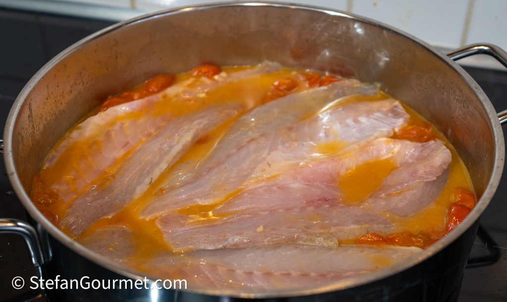 A pot with branzino fillets and halved cherry tomatoes simmering in a light broth.