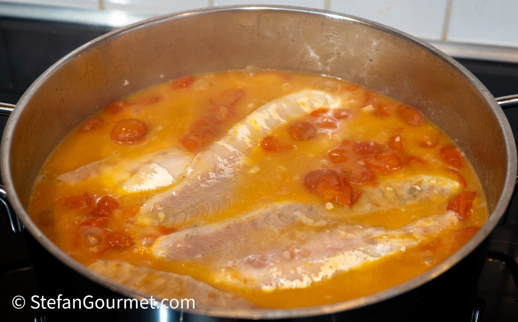 A pot containing branzino fillets poaching in a colorful broth with cherry tomatoes.