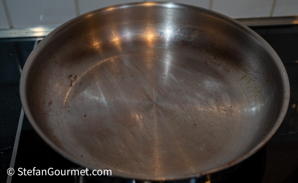An empty stainless steel frying pan with some residual browning, placed on a stovetop.