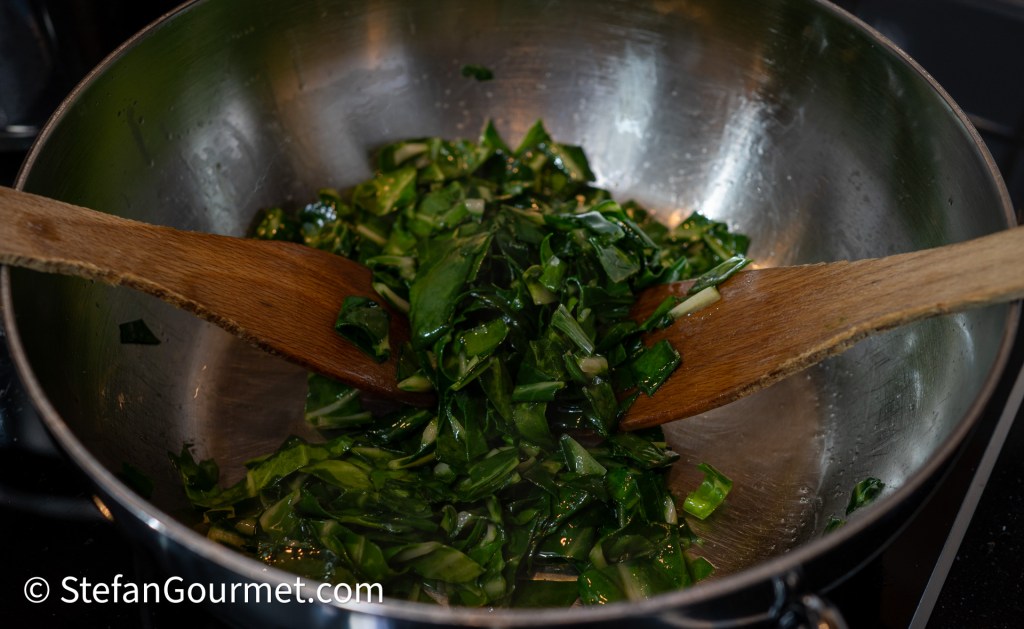 Chopped chard leaves being stir-fried in a stainless steel pan with wooden spoons.
