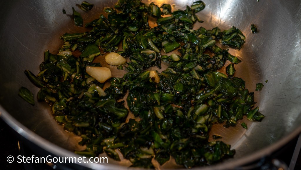 Sautéed chard leaves with garlic in a stainless steel pan, showing a mixture of chopped greens and whole garlic cloves.