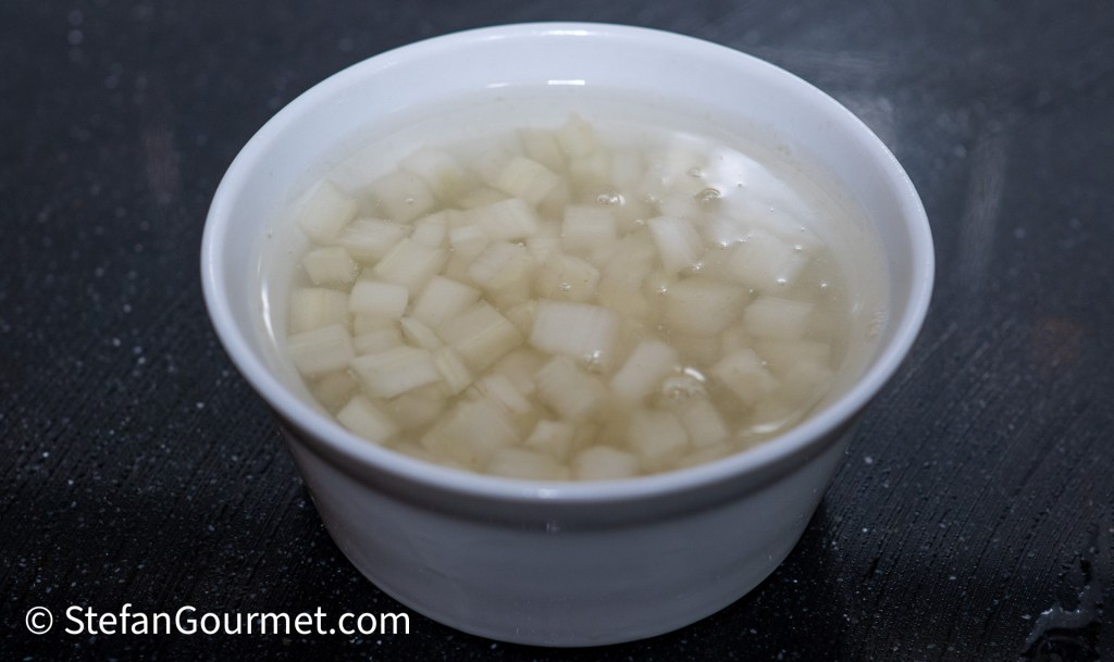 A small white bowl filled with diced parsley root submerged in water, placed on a dark surface.