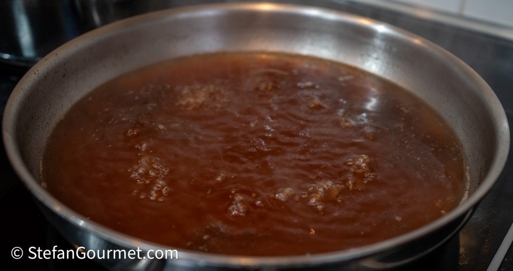 A simmering sauce made from hare bones, dark brown in color, bubbling in a stainless steel pan on a stove.