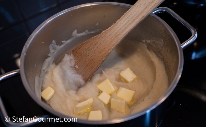 A saucepan on a stovetop containing a creamy mixture, possibly a puree or sauce, being stirred with a wooden spoon, while chunks of butter are melting into it.