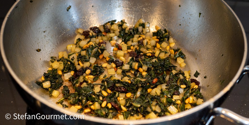 A sauté pan filled with a mixture of chopped chard, diced ingredients, raisins, and pine nuts, all lightly cooked and glistening.