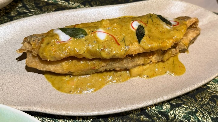 A serving of deep-fried sea bass fillet topped with a rich yellow curry sauce, garnished with red chili and fresh herbs, presented on a decorative plate.