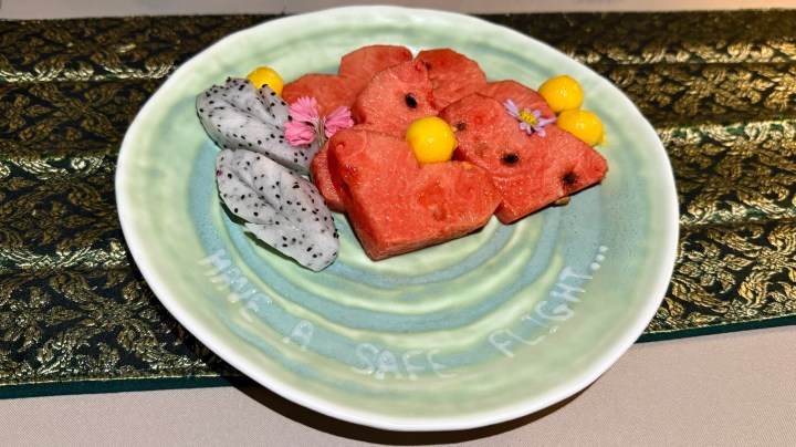 A decorative plate featuring heart-shaped watermelon slices, yellow melon balls, dragon fruit, and edible flowers, with a message written on the plate saying 'Have a safe flight...'.