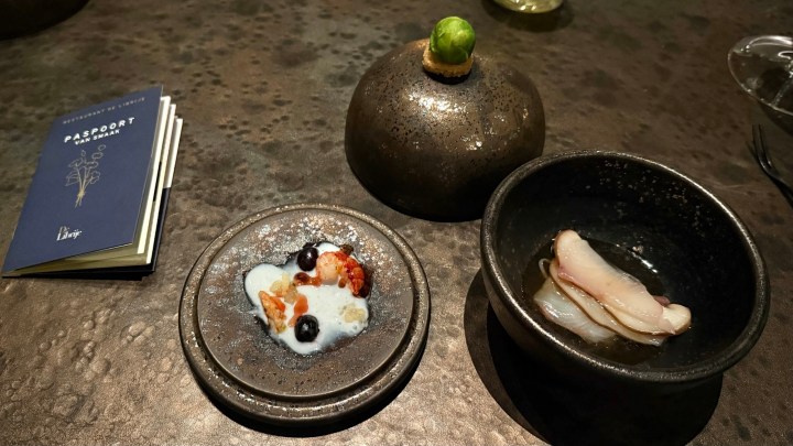 A dish presented at De Librije featuring a small plate with a creamy mixture and colorful toppings, alongside a bowl containing a slice of fish and a covered dish with a green garnish, all arranged on a textured surface.