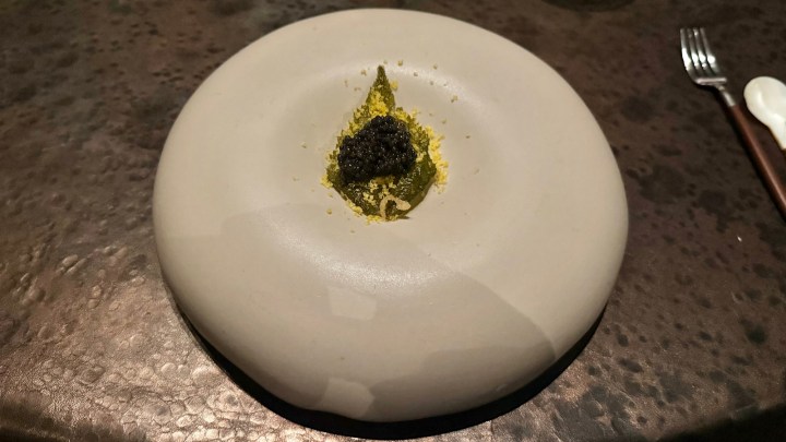 A minimalist dish presented on a round gray plate, featuring a small mound of green puree topped with caviar and finely sprinkled yellow powder.