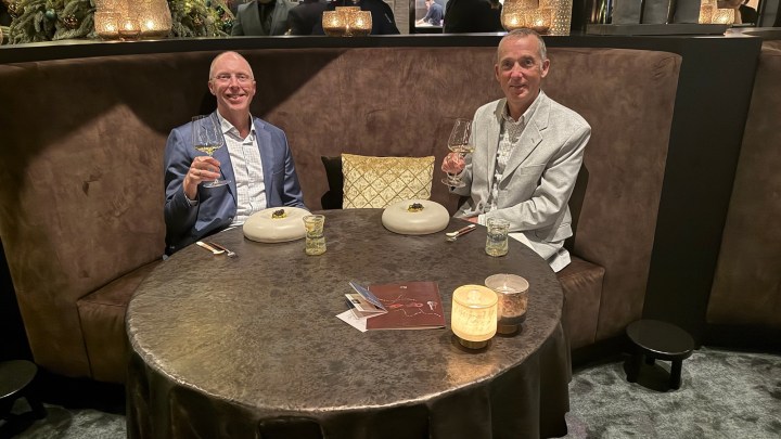Two individuals seated at a round table in an elegant restaurant, each holding a drink and smiling.