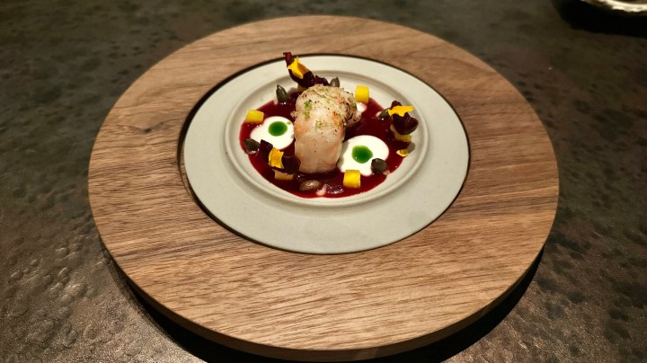 A plated dish featuring a delicate piece of fish garnished with colorful ingredients, including diced mango, dark beans, and green sauce, served on a round wooden plate.