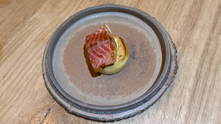 A single bite-sized dish featuring a small round pastry topped with a slice of crispy bacon, served on a textured plate.