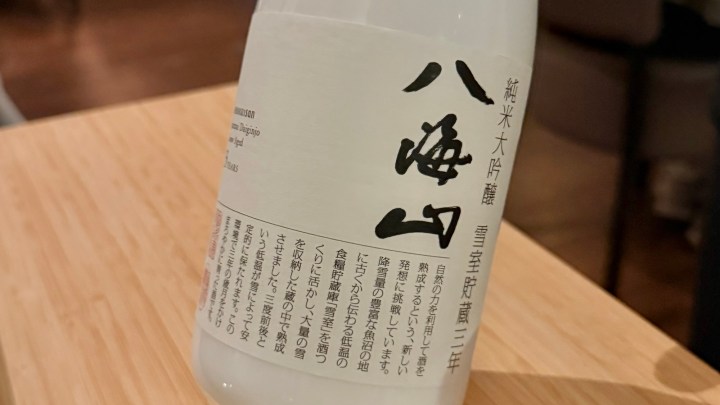 A close-up of a bottle of sake featuring Japanese text on a white label, resting on a wooden table.