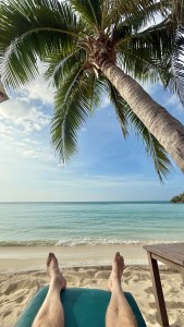 A person's feet resting on a beach chair with a view of the ocean and palm trees in Thailand.