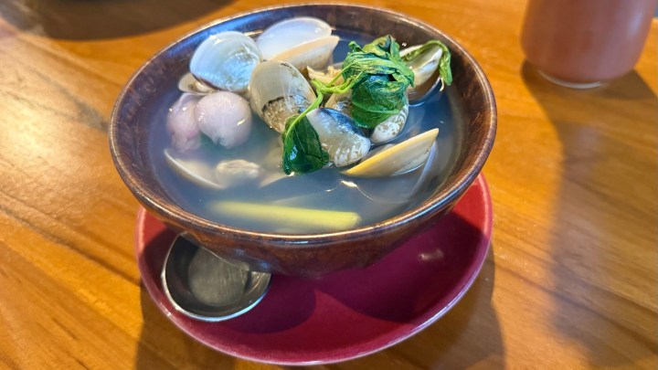 A bowl of clams in a light broth with Thai sweet basil and lemongrass, served on a red plate.