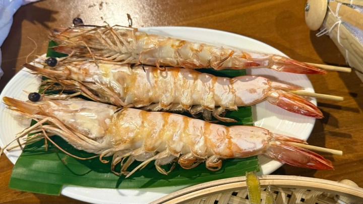 A plate of large grilled prawns skewered for presentation, garnished with banana leaves, placed on a wooden table.