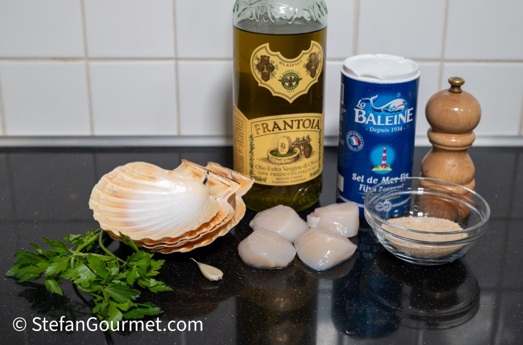 Ingredients for a seafood dish including scallops, a bottle of extra virgin olive oil, sea salt, ground pepper, garlic, parsley, and breadcrumbs on a countertop.