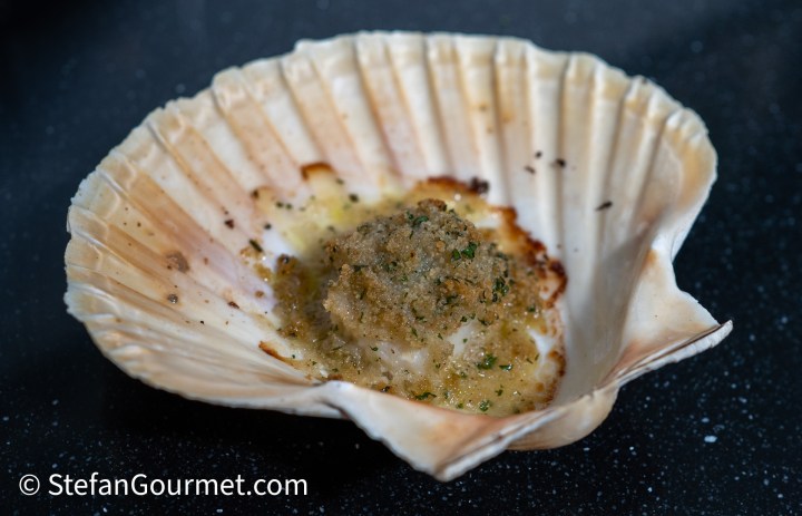 A baked scallop presented in its shell, topped with a golden breadcrumb mixture and sprinkled with herbs, placed on a dark background.